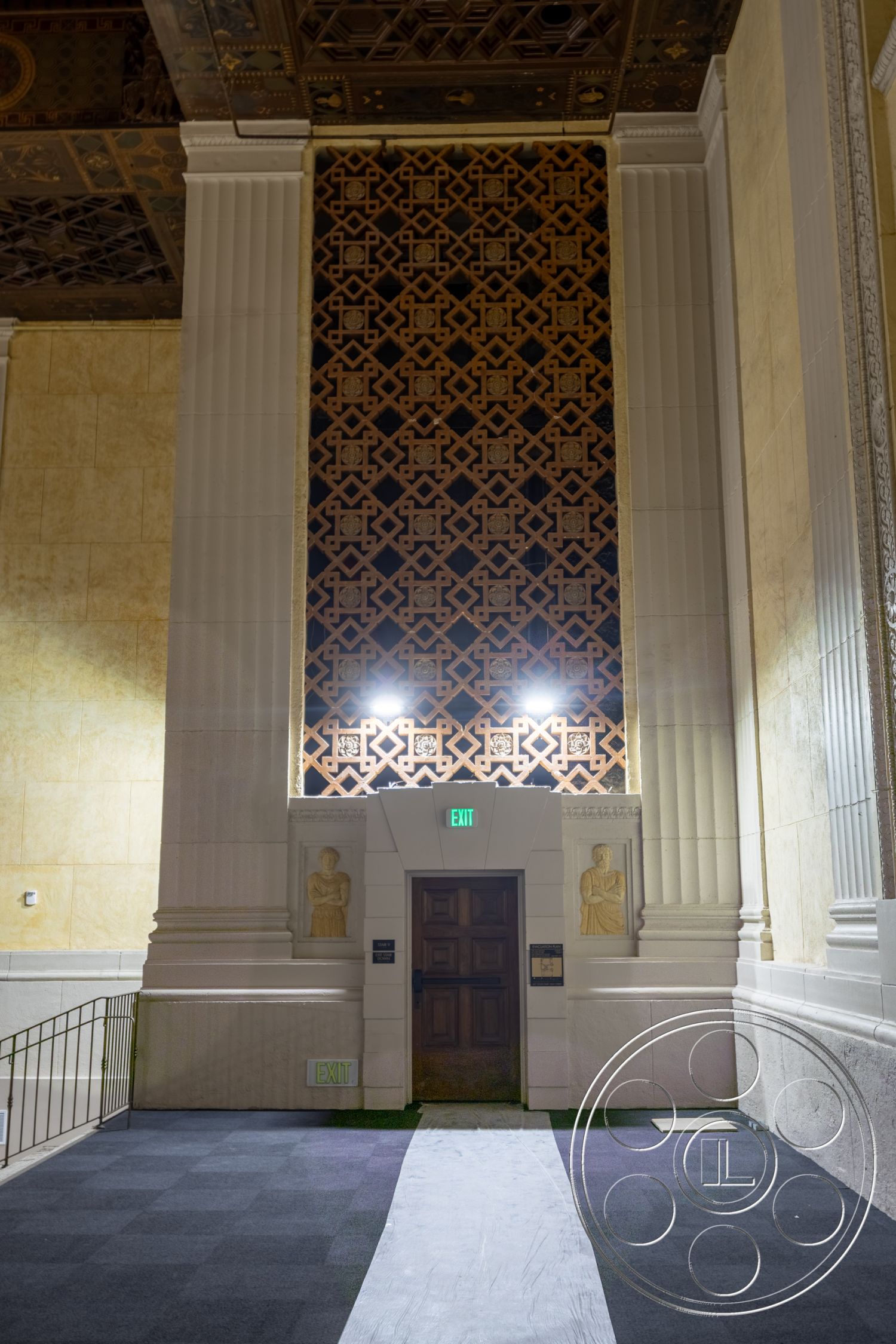 The Plaza - carpet flooring, ornate ceiling details, textured wall treatments, marble accent wall, dark wood door, columned entrance, geometric wall paneling, elaborate crown molding, decorative figures in wall recesses, exposed lit exit sign, high ceiling with plasterwork