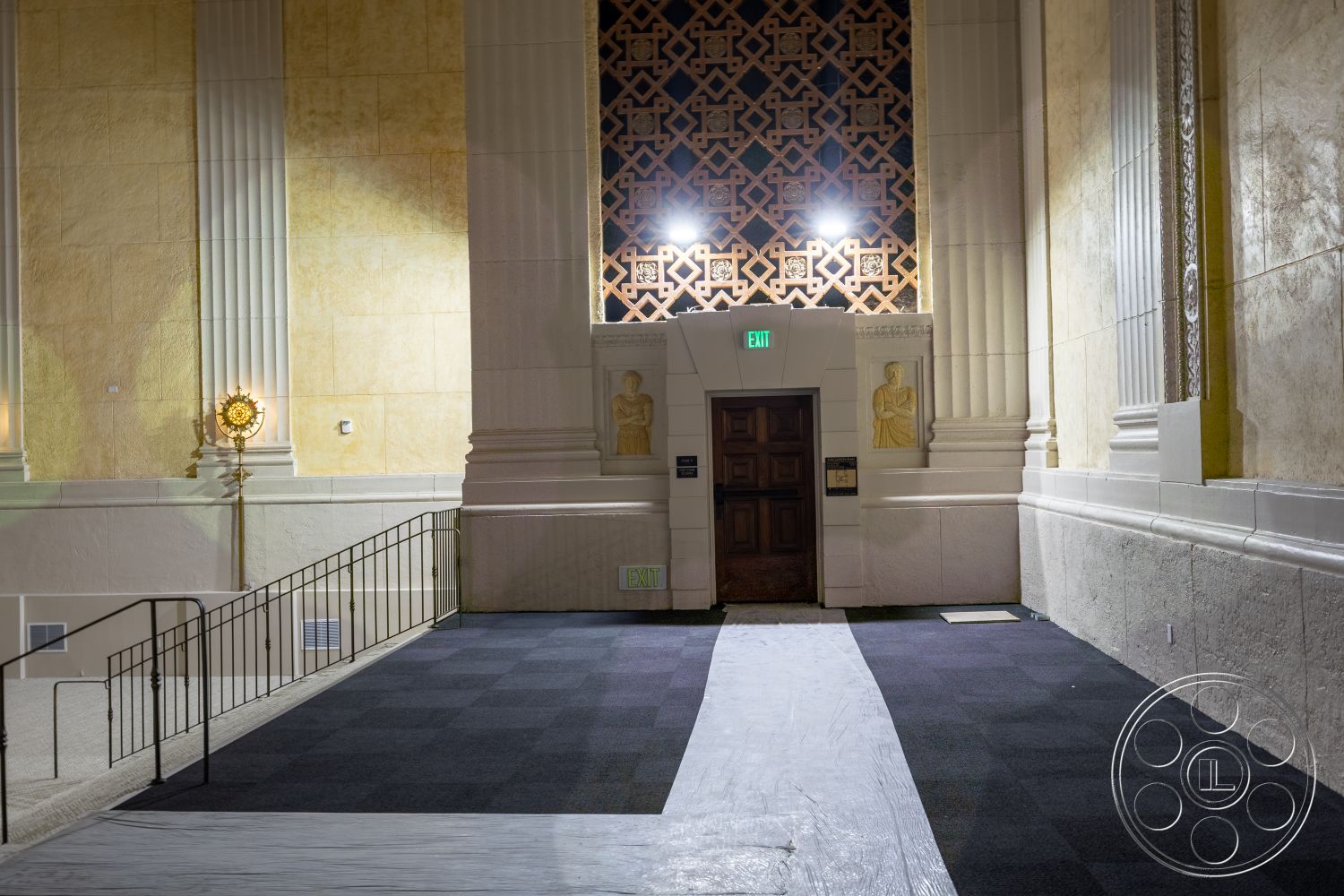 The Plaza - metal railing, carpet flooring, textured plaster walls, ornate ceiling details, dark wood door, decorative wall paneling, column supports, illuminated accent features