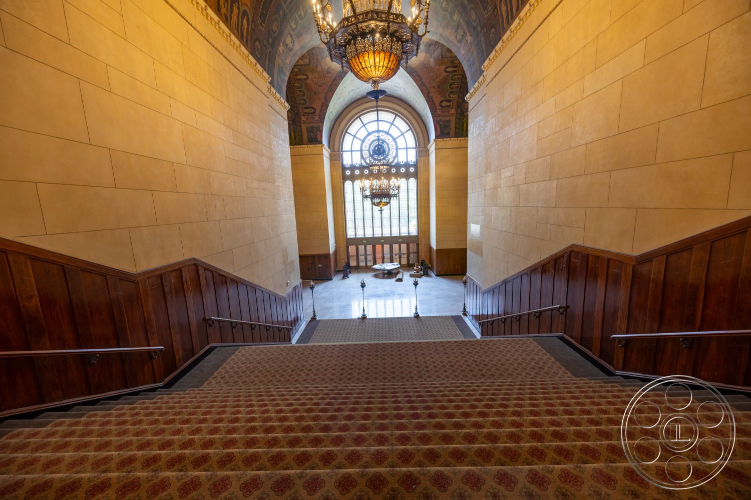 The Plaza - textured wall finishes, historic architectural style, staircase with patterned carpet flooring, wood paneling wall details, vaulted ceiling with decorative mural, large arched window with circular design, intricate chandelier lighting fixture, metal railing on staircase, marble floor at landing area