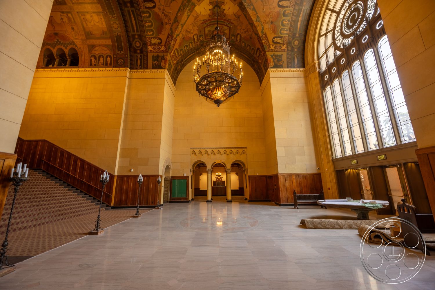The Plaza - high vaulted ceiling, marble flooring, arched doorways, large arched windows, ornate chandelier, grand hall, staircase with metal railings, historic architectural style, wood paneling details, exposed stone walls, decorative ceiling murals, entry foyer