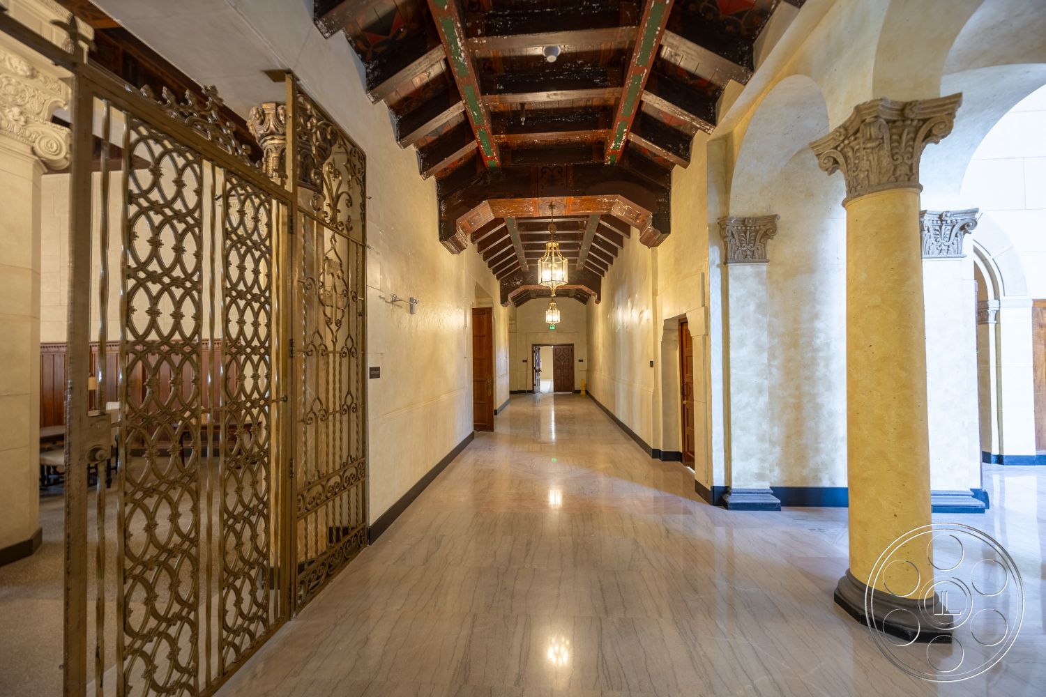 The Plaza - marble flooring, arched doorways, dark wood paneling, brass light fixtures, exposed wood ceiling beams, coffered wood ceiling, painted plaster walls, gold ornamental iron gate, detailed column capitals, clear glass pendant lights