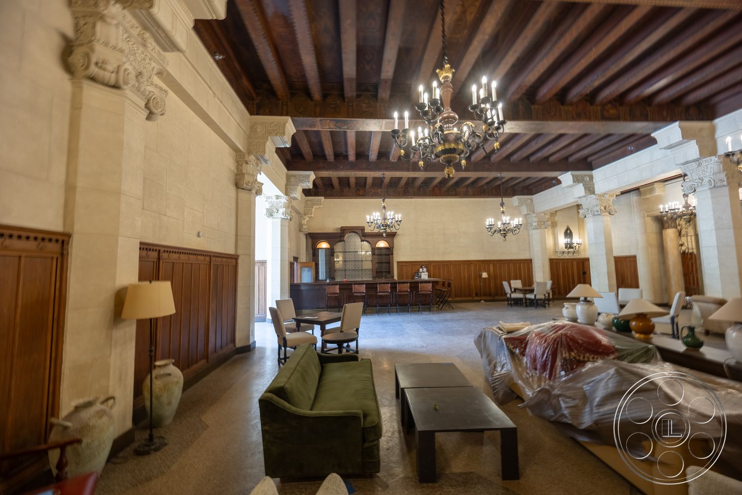The Plaza - modern coffee table, polished stone flooring, dark wood paneling, exposed wood ceiling beams, intricate crown molding details, spacious open floor plan, ornate chandelier lighting, marble accent columns, carved stone wall panels, traditional style furnishings, central bar area with wooden counter, classic seating arrangement, large decorative vases