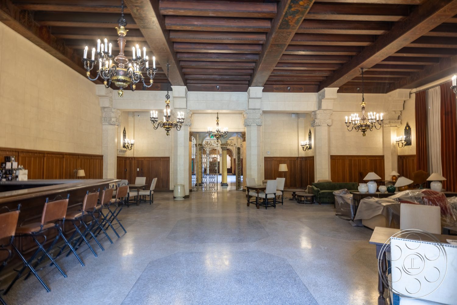 The Plaza - neutral color palette, arched doorways, dark wood paneling, elegant lounge area, ornate wall sconces, exposed wood ceiling beams, decorative molding details, chandelier lighting fixtures, traditional seating area, stone wall panels, smooth polished marble flooring, long bar counter, ceiling medallions