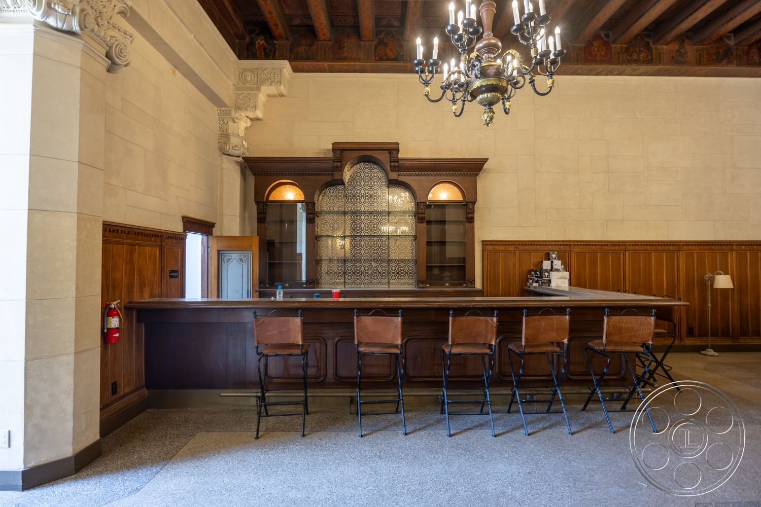 The Plaza - stone accent wall, arched doorway, dark wood paneling, ornate ceiling details, intricate chandelier, exposed wood ceiling beams, wood bar countertop, tile bar back wall, marble bar floor, vintage metal bar stools, built-in glass display cabinets