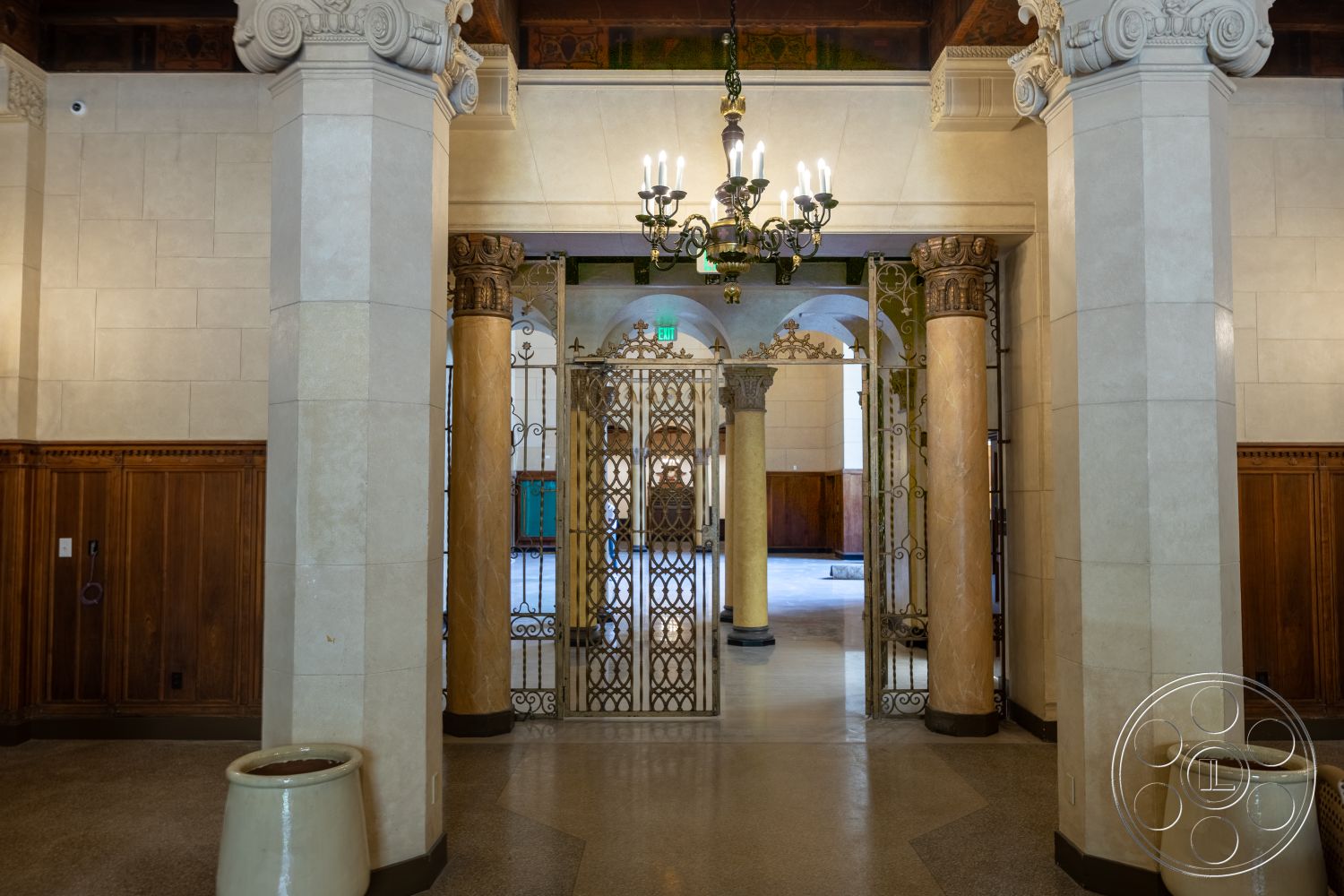 The Plaza - exterior, arched entryway, dark wood paneling, smooth tile flooring, coffered ceiling details, textured stone wall panels, chandelier lighting fixture, decorative wrought iron gates, marble column bases, ornate plaster ceilings