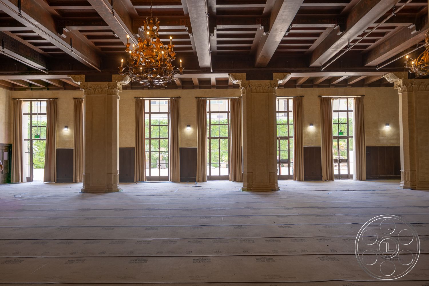 The Plaza - textured plaster walls, large floor-to-ceiling windows, exposed wood ceiling beams, chandelier lighting fixture, wood paneling details, curtained window treatments, stone column bases, unfinished flooring