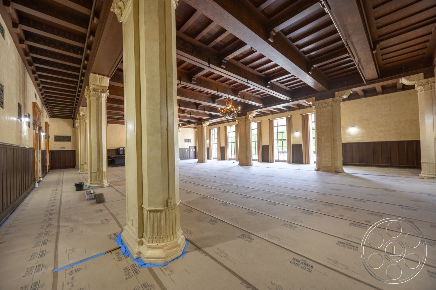 The Plaza - natural lighting, dark wood paneling, large floor-to-ceiling windows, classic architectural style, exposed wooden ceiling beams, open layout space, chandelier lighting fixture, plastered wall finish, ornate column details, stone column bases, cream-colored flooring substrate, white painted wall accents