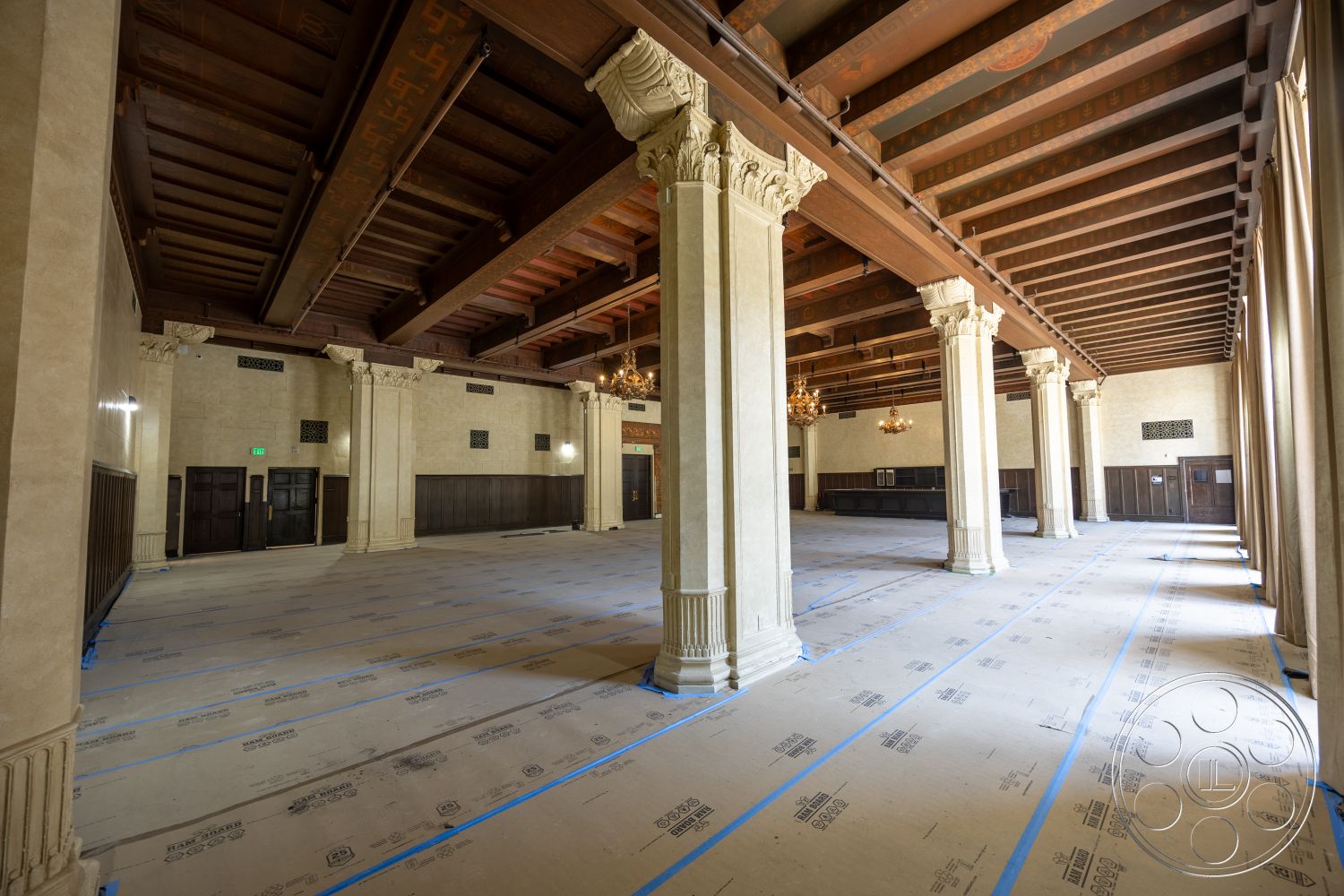 The Plaza - wood paneling, large open space, vaulted ceiling, chandelier lighting, plastered walls, ornate ceiling details, unfinished concrete floor, classic architectural style, exposed wood ceiling beams, fluted columns, symmetrical room layout, column support structure, historic venue