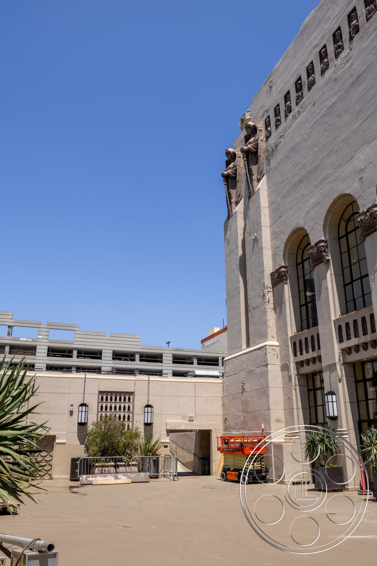 The Plaza - exterior, concrete walls, landscaping with tropical plants, historical building facade, circular entryway, symmetrical building design, windowed arches, decorative wall reliefs, art deco architectural details, outdoor plaza area, ground-level courtyard, industrial lifting equipment, light fixtures with art deco styling