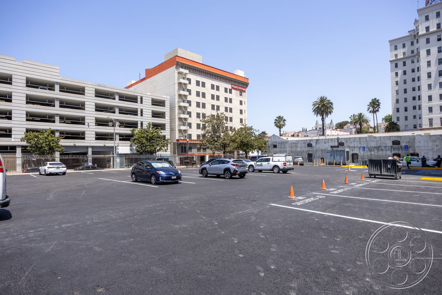 The Plaza - exterior, asphalt surface, urban setting, accessible parking, striped parking spaces, concrete parking lot, flat roof design, city background, modern industrial architecture, painted concrete walls, concrete parking structure, palm trees lining the area, multiple parking levels, recessed building entrance