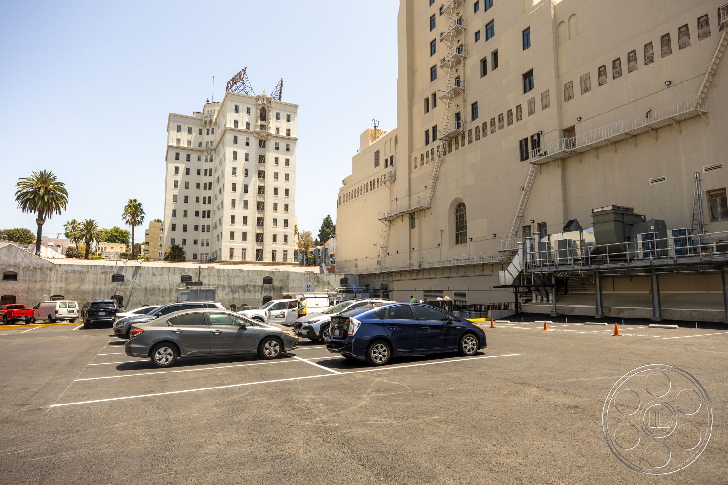 The Plaza - large windows, exterior, white stucco walls, multi-story building, modernist architectural style, palm trees in urban setting, concrete wall perimeter, parking lot asphalt surface, steel fire escape stairs, industrial air conditioning units