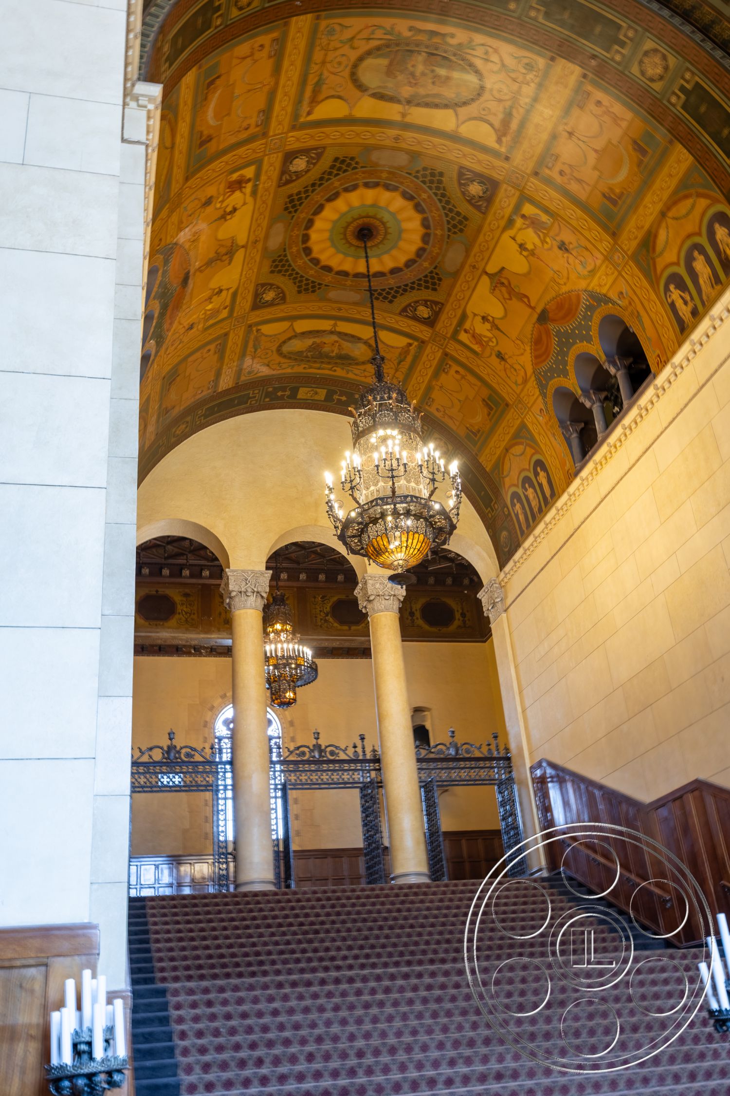 The Plaza - columned support structure, chandelier lighting fixtures, decorative wrought iron railings, ornate ceiling with decorative murals, exposed wooden handrails, carpeted stairs with intricate pattern, tall arched doorways, polished stone wall panels, vaulted grand entrance hall