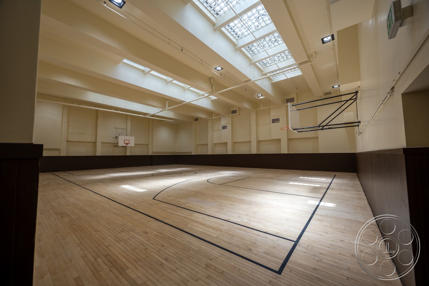 The Plaza - open space layout, high vaulted ceiling, bright overhead lighting, dark wood wainscoting, maple hardwood basketball court flooring, curved gymnasium wall, skylights with glass panels, basketball hoop fixture, wooden court markings