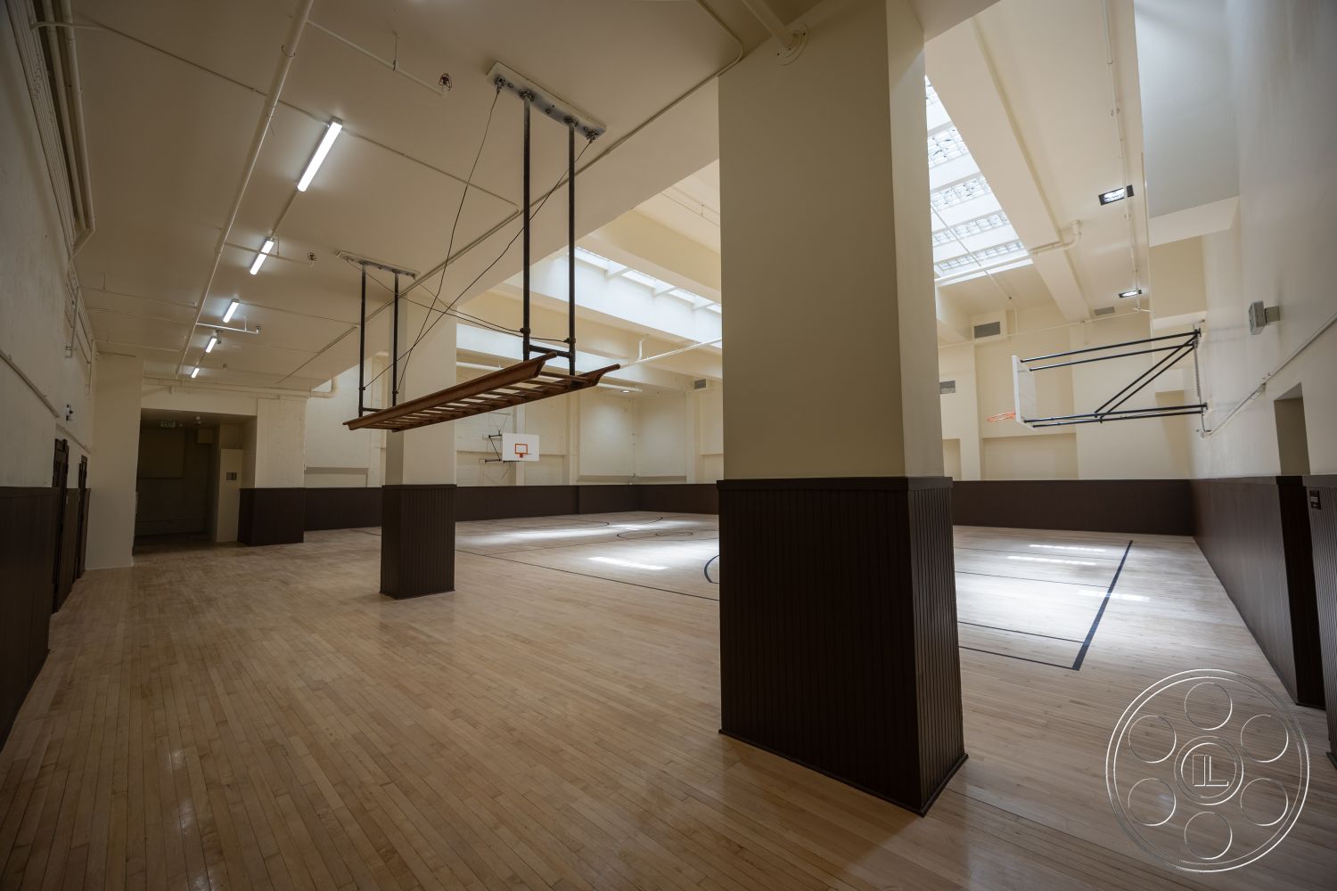 The Plaza - brown wood paneling, exposed ceiling structure, white painted walls, natural light from skylights, high ceiling with skylights, hardwood gymnasium flooring, basketball court with markings, overhead basketball hoop, open layout gym area