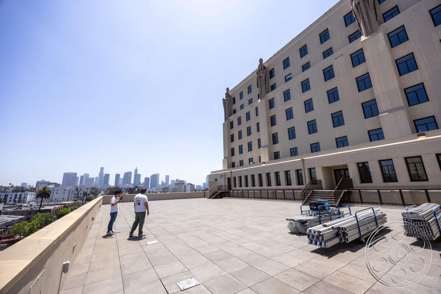 The Plaza - exterior, urban environment, steel railing, modern architectural style, flat roof design, large rectangular windows, high-rise city view, concrete building facade, plaza terrace with stone tile flooring, terrace overlooking city skyline, decorative stone statues at corners, minimalist outdoor furniture arrangement