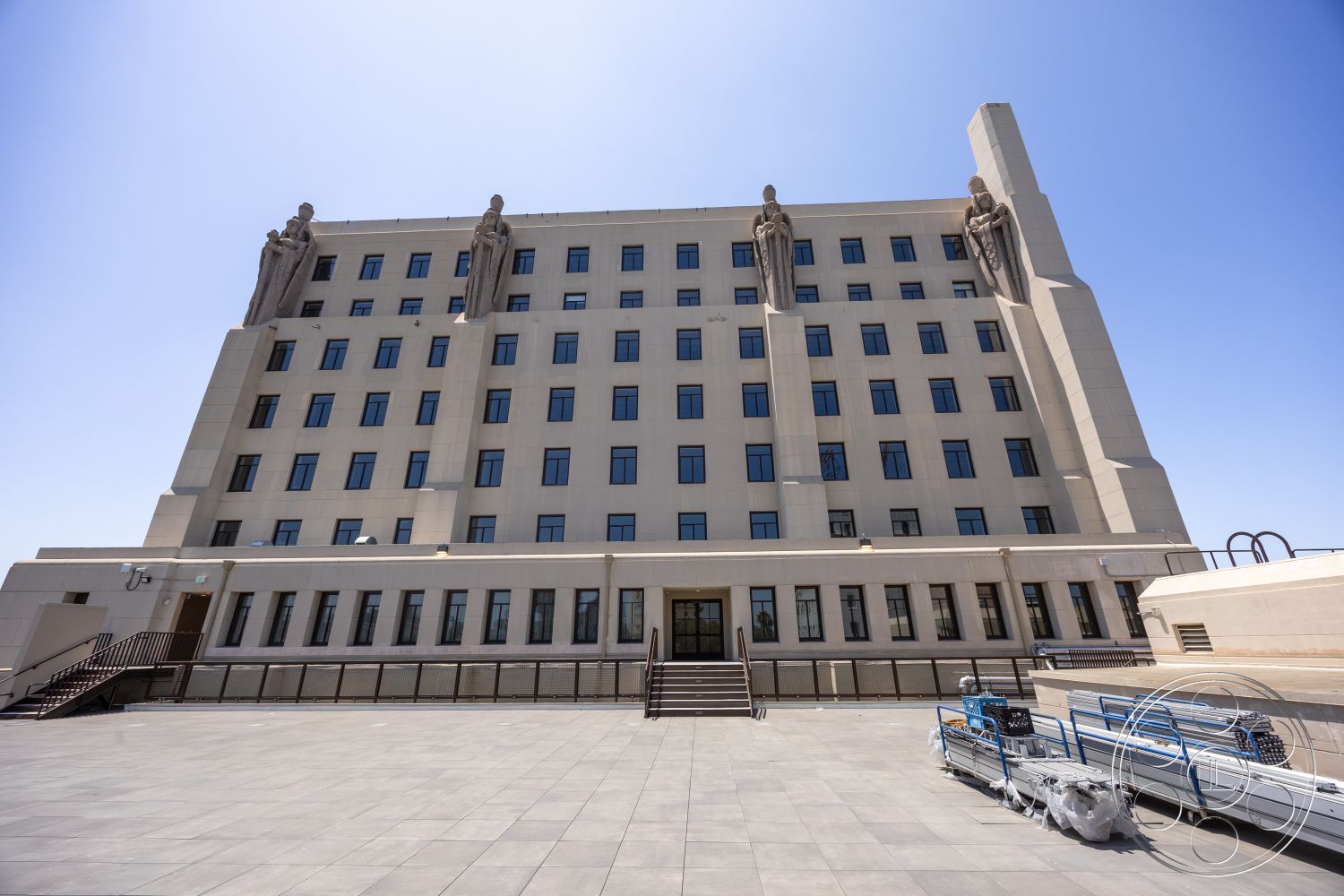 The Plaza - exterior, flat roof, metal railings, large staircase, rectangular windows, modernist architectural style, concrete building facade, geometric architectural details, stylized stone sculptures, plaza paving stones, public building entrance