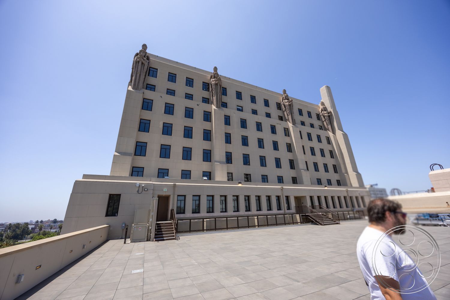 The Plaza - exterior, flat roof, urban setting, commercial building, stone facade, rectangular windows, large outdoor terrace, steel stair railings, symmetrical building design, concrete paving stones, sculptural architectural details, art deco style elements, statues flanking the building, horizontal banding on walls