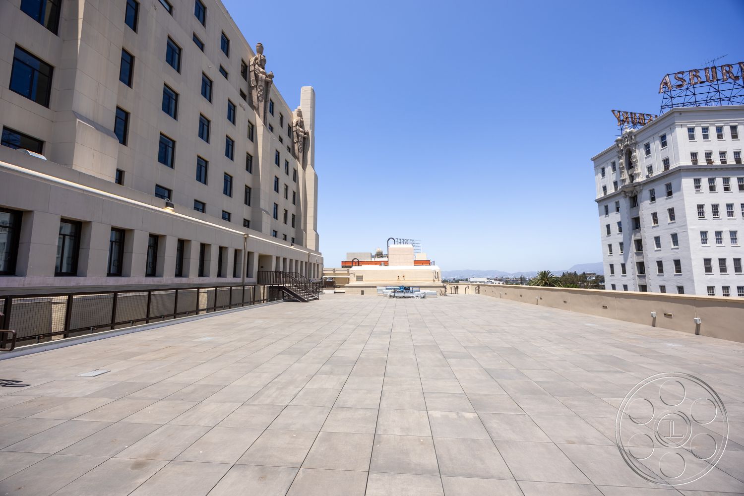 The Plaza - exterior, city skyline view, flat roof design, large rectangular windows, concrete terrace flooring, historical architectural style, commercial building environment, metal railing perimeter, rectangular rooftop layout, modern urban building facade, brick wall details, prominent architectural sculptures