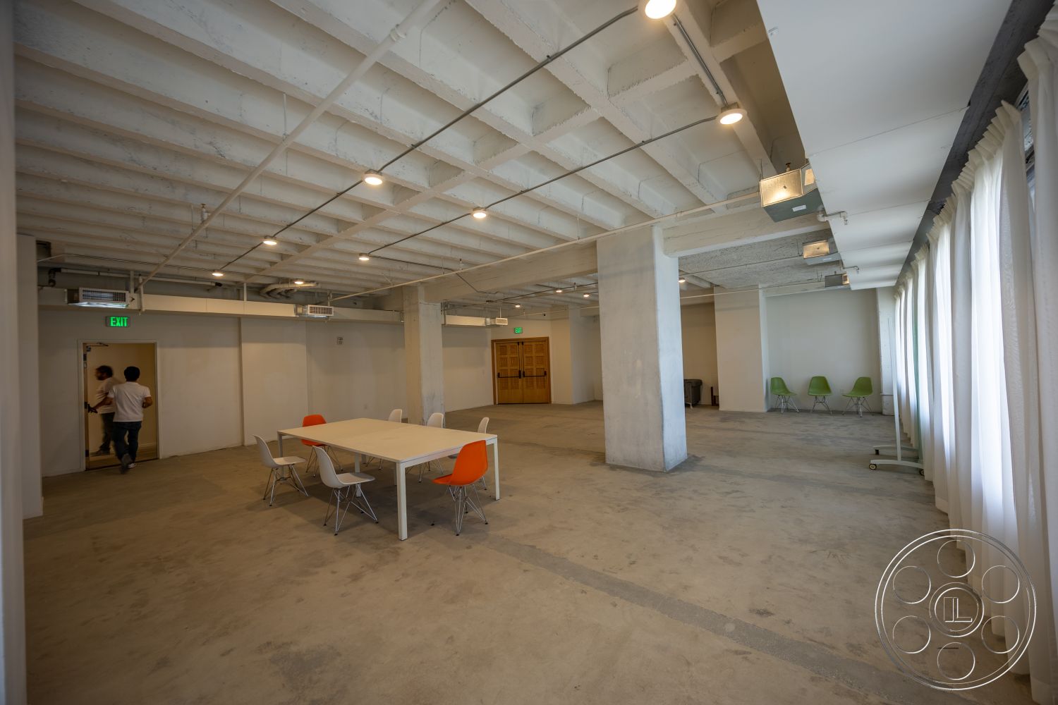 The Plaza - neutral color palette, open floor plan, concrete floor, modern workspace, floor-to-ceiling windows, industrial lighting fixtures, exposed concrete ceiling, orange accent chairs, metal dining chairs, white painted walls, rectangular conference table, soundproof curtain panels