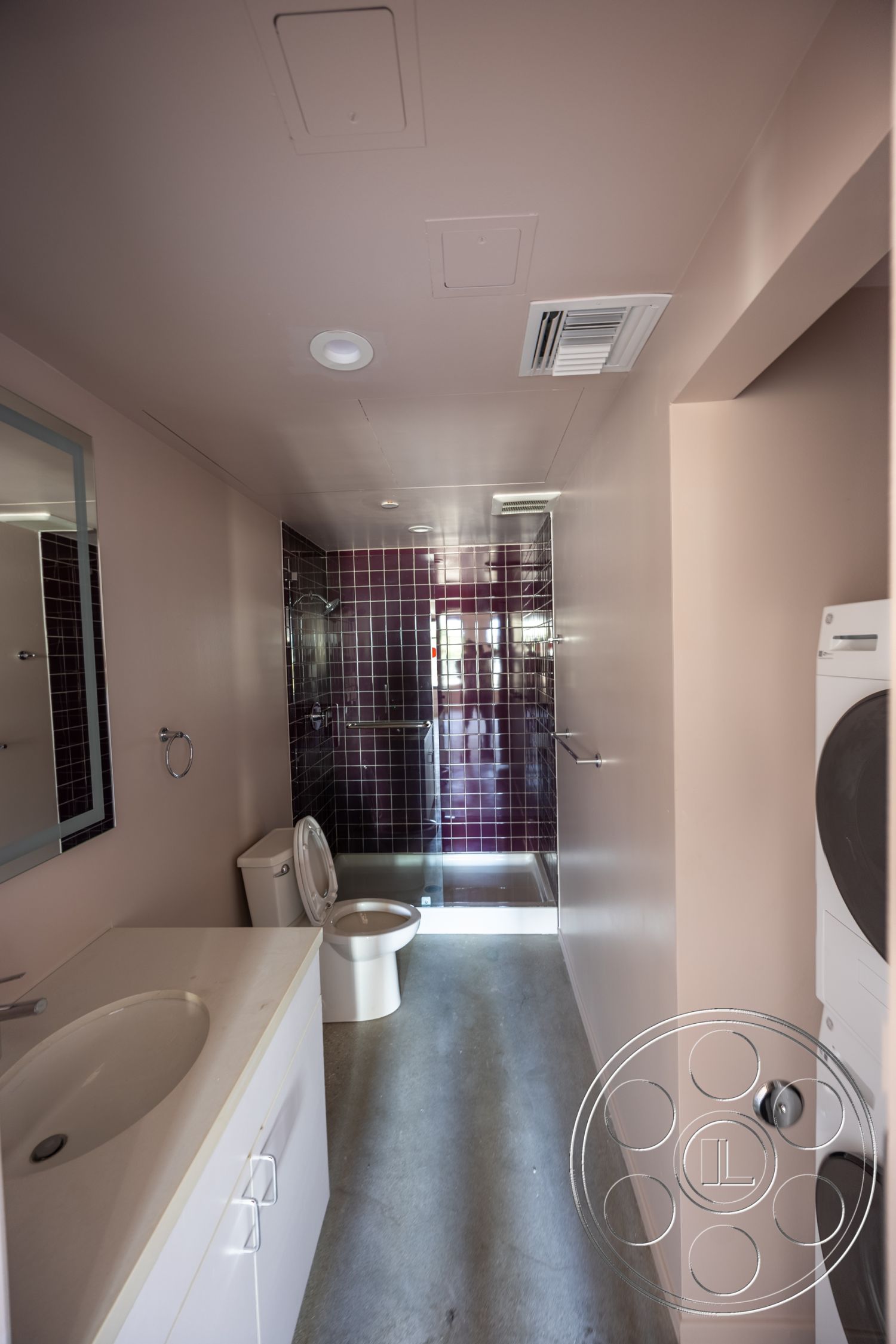 The Plaza - glass shower enclosure, polished concrete flooring, modern lighting fixtures, plastered walls, interior bathroom, white vanity cabinet, white sink countertop, purple tile shower walls, round toilet, storage space for laundry appliances