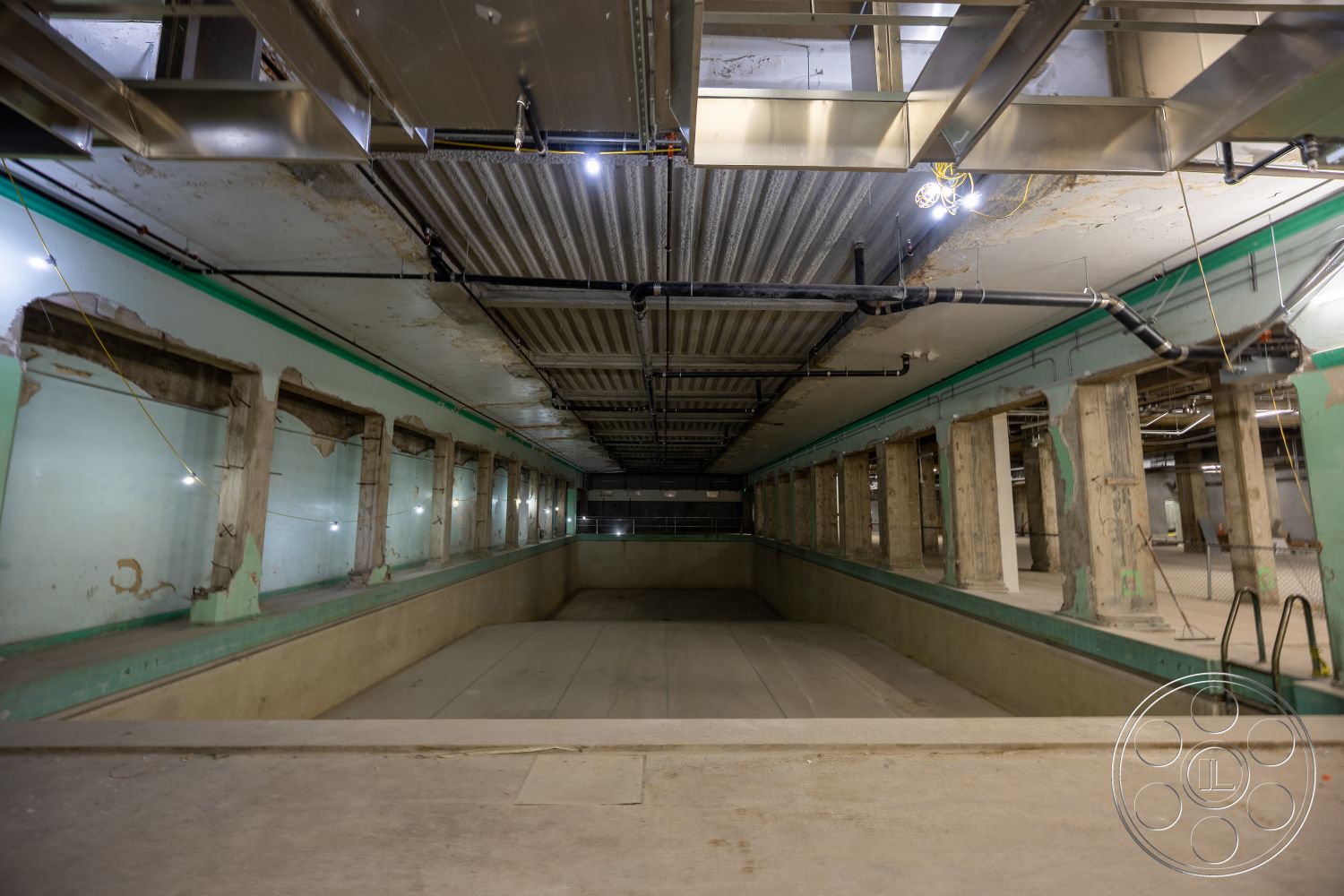 The Plaza - exposed concrete walls, industrial lighting fixtures, steel support beams, glass block windows, metal roofing panels, warehouse-style architecture, open ceiling with ductwork, concrete pool basin, unfinished floor surface, painted green accents