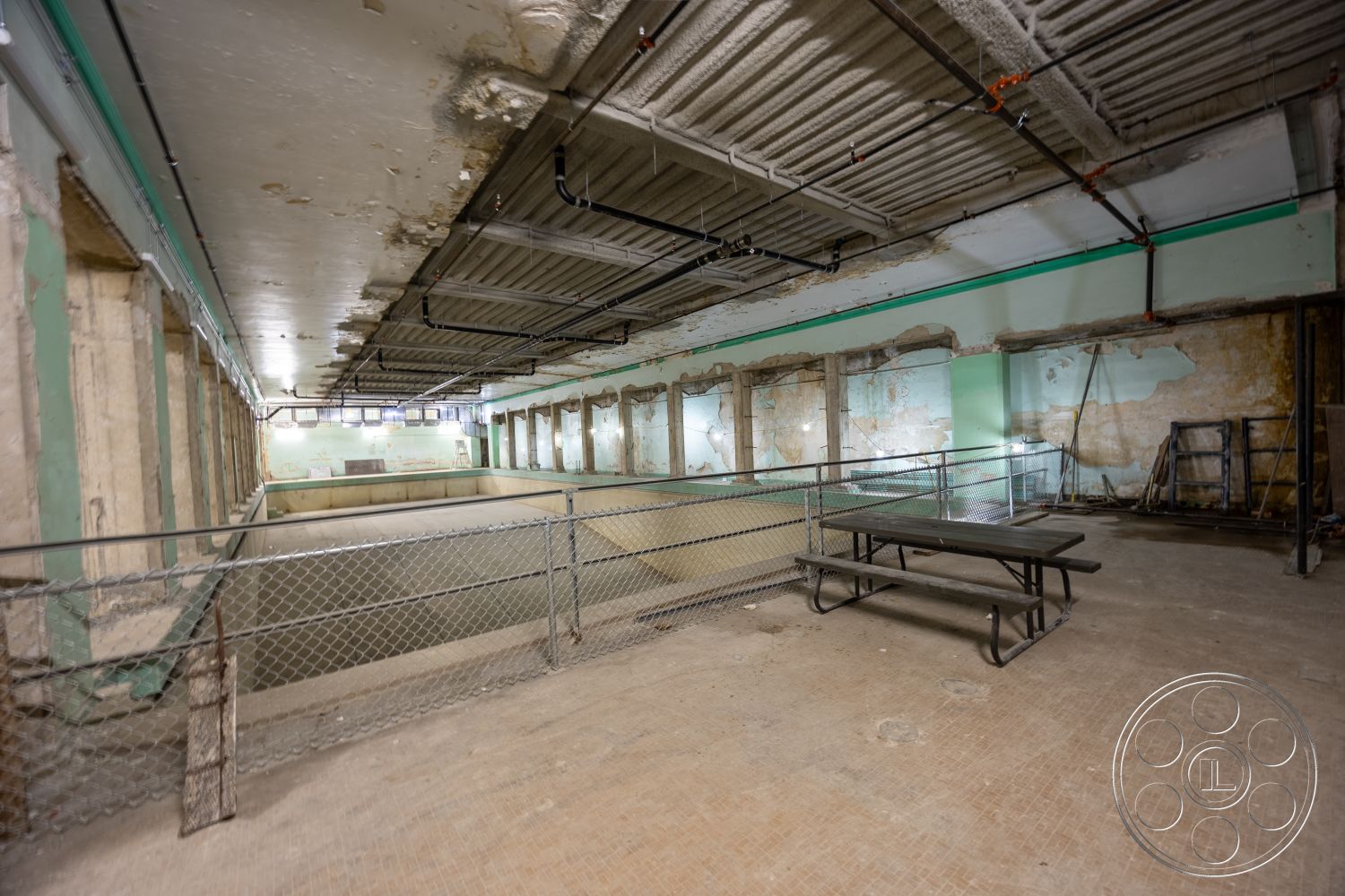 The Plaza - large windows, open floor plan, interior space, exposed concrete walls, industrial style, unfinished ceiling, multipurpose area, sparse furniture, metal ceiling beams, chain link fencing, green painted accent wall, plaster wall remnants, remnants of tiled flooring, plumbing fixtures visible, light fixtures mounted on ceiling, former swimming pool area