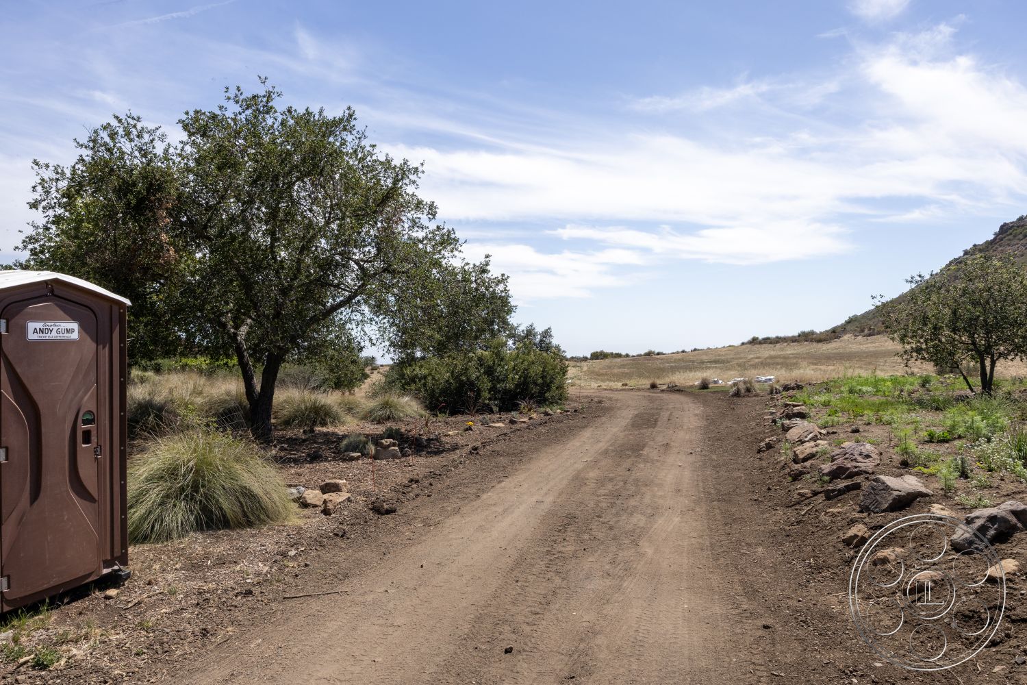 Eagle Feather - gravel driveway, mountainous backdrop, dirt road, portable restroom, outdoor environment, natural rock border, native vegetation landscape, open grassy field setting