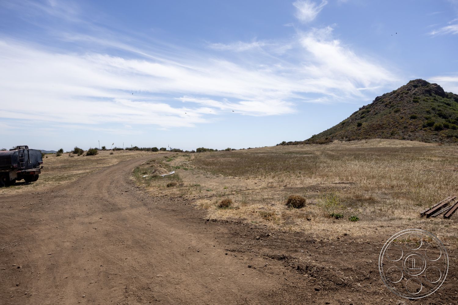 Eagle Feather - exterior, clear blue sky, natural setting, dirt road, rocky hillside, mountainous terrain, rural landscape, dry grass landscape, vegetation patches, scattered clouds, deserted environment