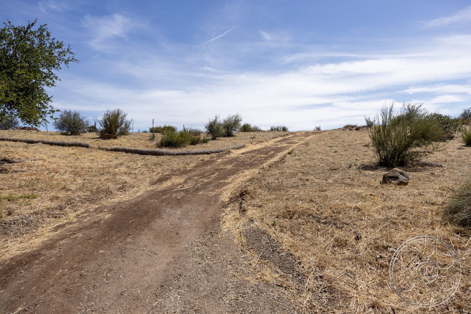 Eagle Feather - exterior, hillside setting, rocky terrain, scattered shrubs, blue sky with clouds, dirt access road, dry grassy landscape