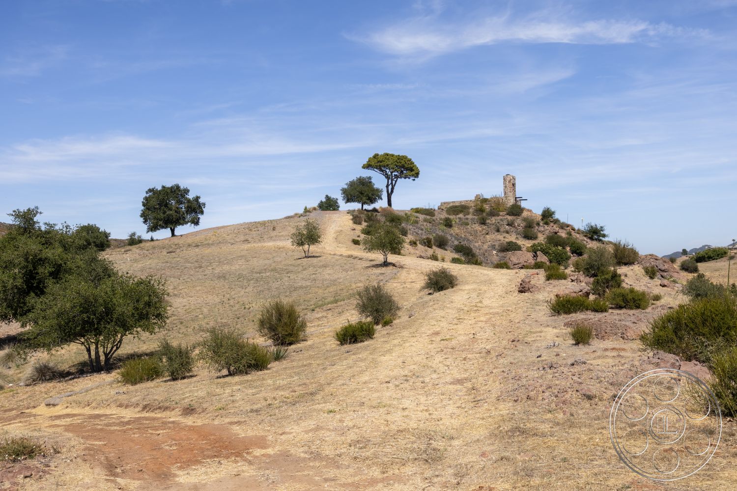 Eagle Feather - natural landscape, blue sky, rocky hillside, scattered trees, sunny environment, pathway leading uphill, dry grass terrain, distant ruins, vegetation with shrubs