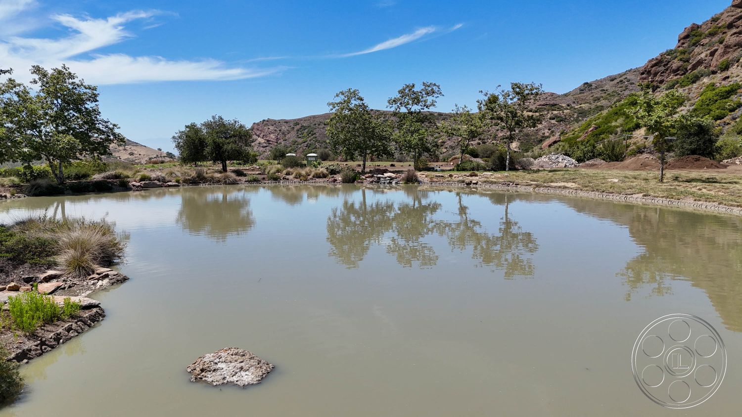 Eagle Feather - exterior, clear blue sky, dirt pathway, tall grass, mountainous background, surrounding hillside landscape, open rural setting, natural pond, rocky pond border, reflected tree silhouettes, shrubs and greenery