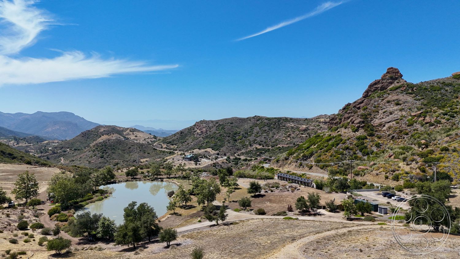 Eagle Feather - exterior, open space, natural vegetation, natural landscape, rural setting, dirt pathway, mountainous terrain, scenic hillside view, distant mountains, lush green trees, agricultural land, calm pond surface, rocky hillside formations, grassy meadows, blue sky with wispy clouds