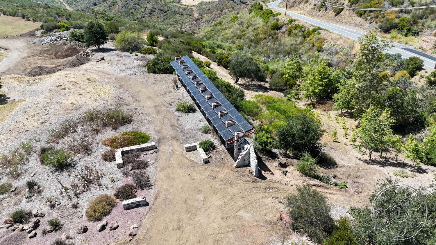Eagle Feather - exterior, natural vegetation, gravel driveway, stone retaining wall, agricultural setting, flat roofing, horizontal wooden slats, solar panel array, open hillside location, scenic mountain backdrop, earth berm landscaping, pathway with native plants