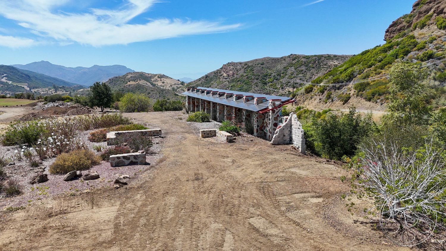 Eagle Feather - exterior, dirt pathway, hillside location, desert vegetation, flat roof design, wood beam roof overhang, landscaped garden beds, concrete foundation, mountainous terrain setting, stone wall construction, rough-hewn stone features, complementary natural landscape