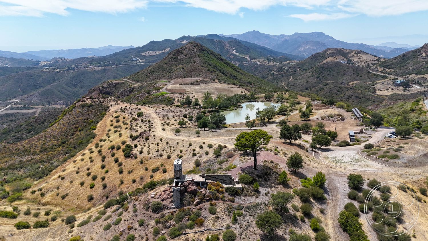 Eagle Feather - exterior, aerial view, mountain retreat, scattered trees, dry grassland, natural landscape setting, desert-like environment, dirt access road, rocky outcroppings, stone tower structure, pond surrounded by greenery, sloping hillside terrain