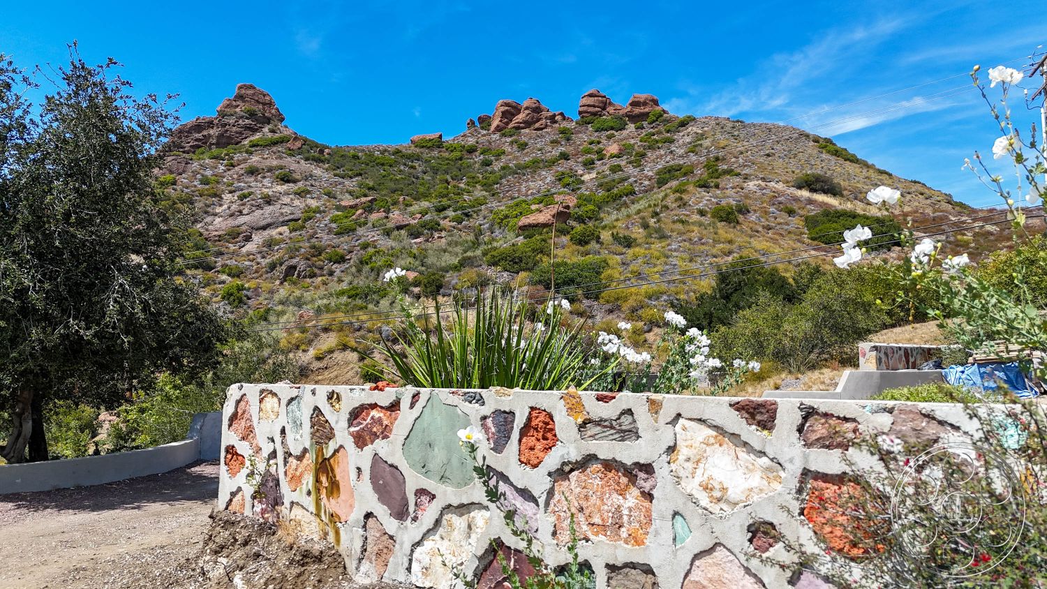Eagle Feather - exterior, lush greenery, flowering shrubs, blue sky, natural stone wall, irregular shaped stone accents, desert hillside setting, cement base, rocky mountain backdrop