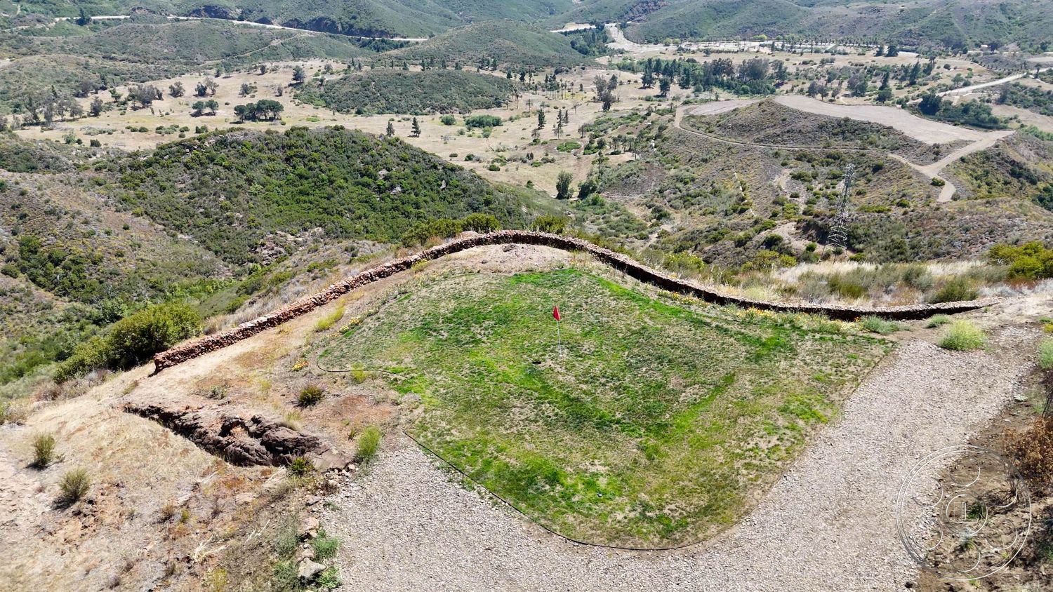 Eagle Feather - exterior, gravel pathway, mountainous backdrop, open landscape, natural stone retaining wall, scenic viewpoint, minimal vegetation, sunlit hillside, curved landscaping border, flat grassy area, surrounding hilly terrain, wooded hillside