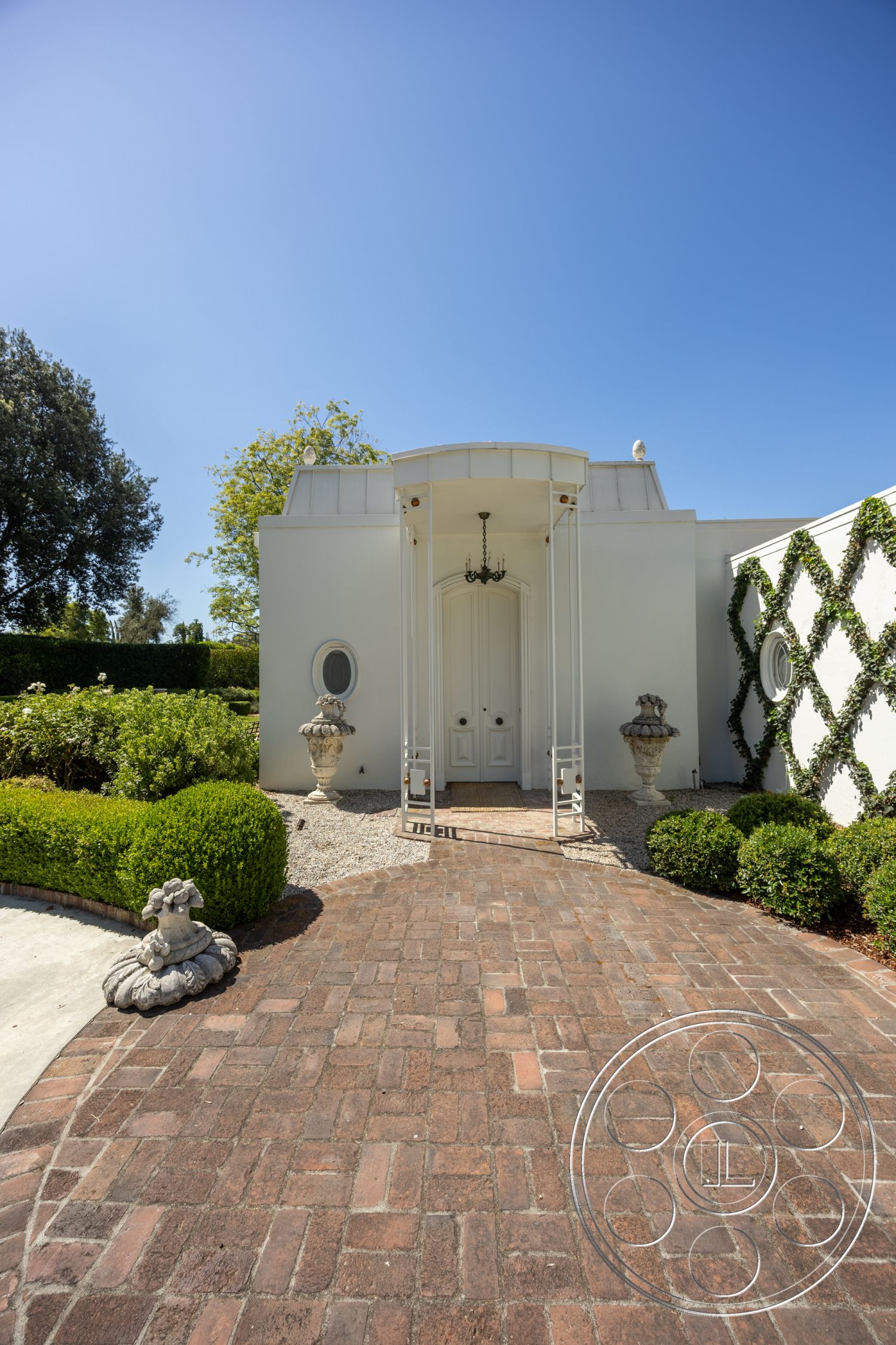 Retro 295 - exterior, curved driveway, arched doorways, white exterior walls, modern residential architecture, landscaped garden area, brick driveway pavers, rounded shrubbery, ornamental stone planters, oval entryway chandelier, textured stucco facade