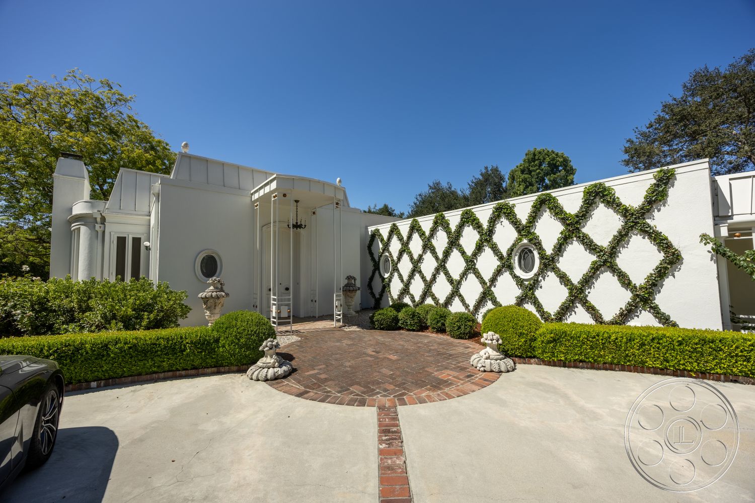 Retro 295 - exterior, white stucco walls, circular driveway, modern architectural style, symmetrical facade, arched entryway, trimmed hedges, landscaped garden beds, brick paver entrance path, geometric lattice trellis, round window accents, ornate exterior lighting fixture