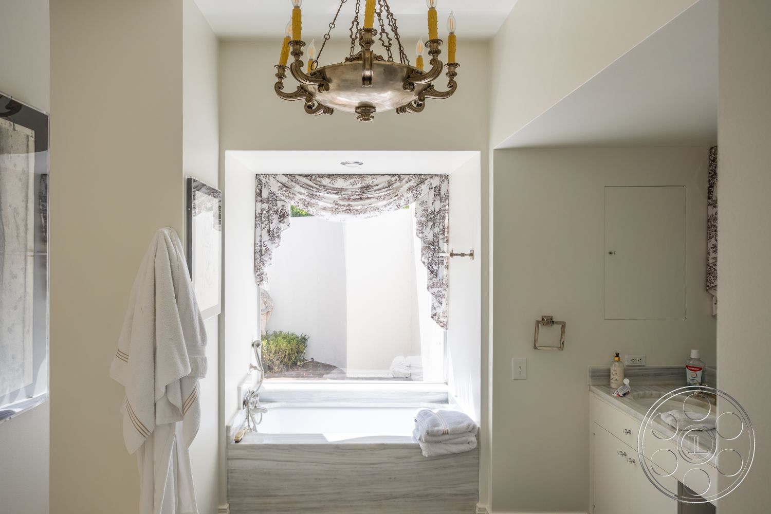 Retro 295 - wood cabinetry, white painted walls, chandelier lighting fixture, ceramic tile bathroom flooring, marble bathtub surround, textured fabric shower curtain, bath towels on countertop, window with fabric treatment, vanity sink area