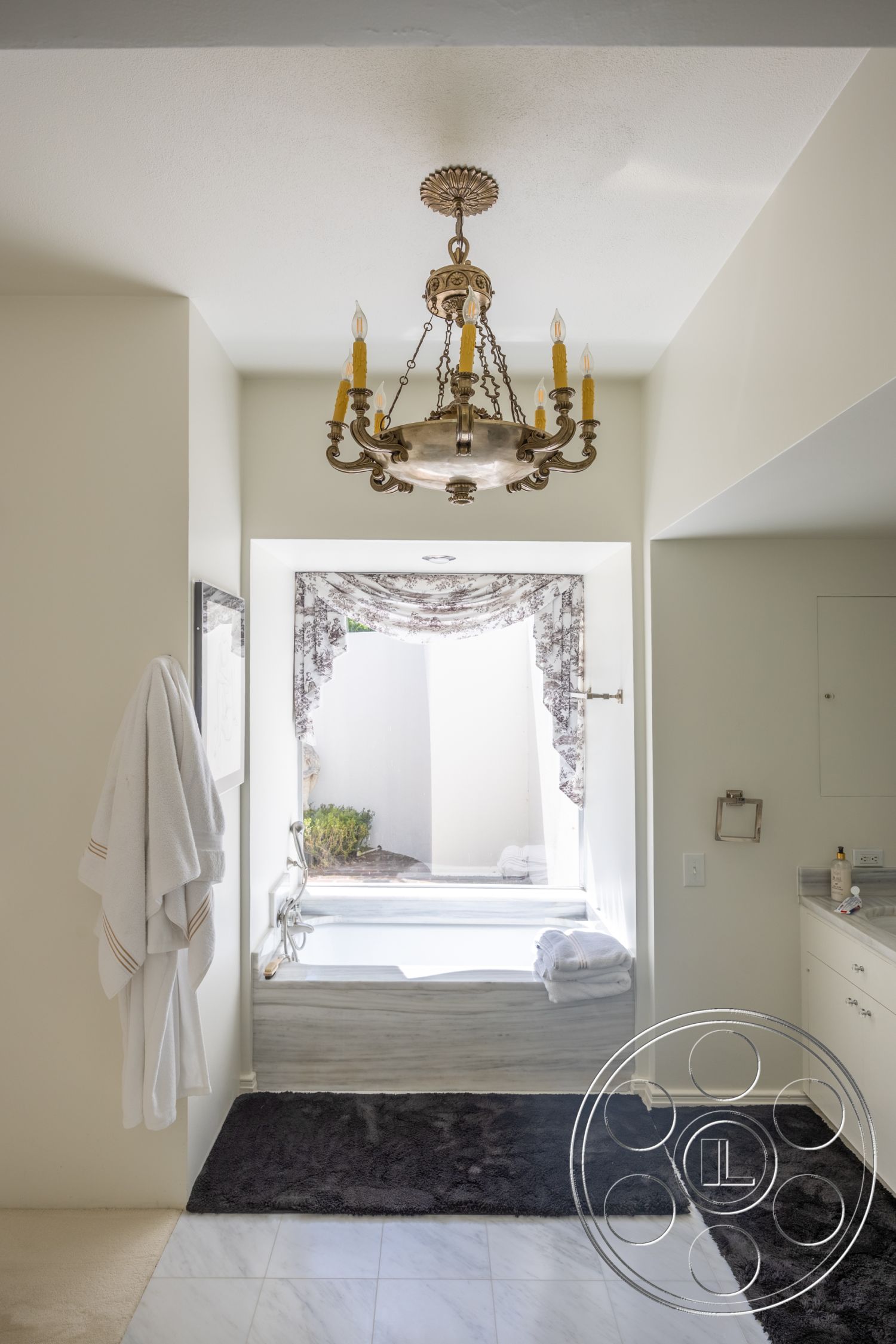 Retro 295 - floor-to-ceiling window, white painted walls, marble bathroom flooring, beige carpet flooring, chandelier lighting fixture, marble bathtub surround, built-in bathroom cabinets, gray plush bath mat, drapery window treatment