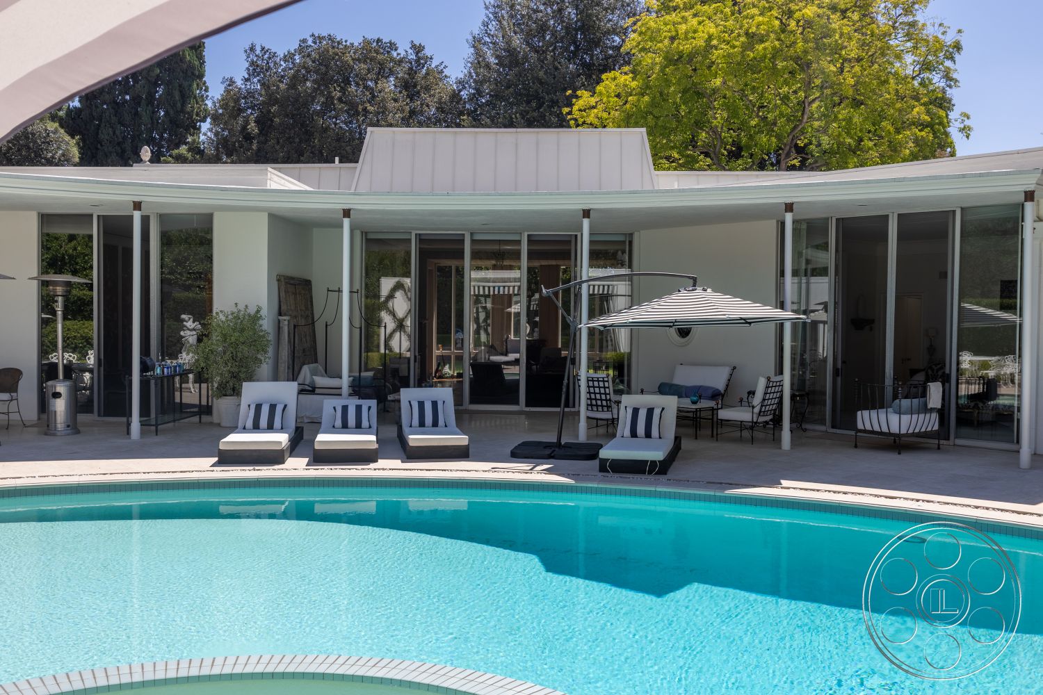 Retro 295 - exterior, white stucco walls, glass sliding doors, outdoor seating area, landscaped greenery, modern architectural style, concrete pool deck, metal roof, curved pool, sun loungers with striped cushions, overhead patio umbrella, patio heating lamp