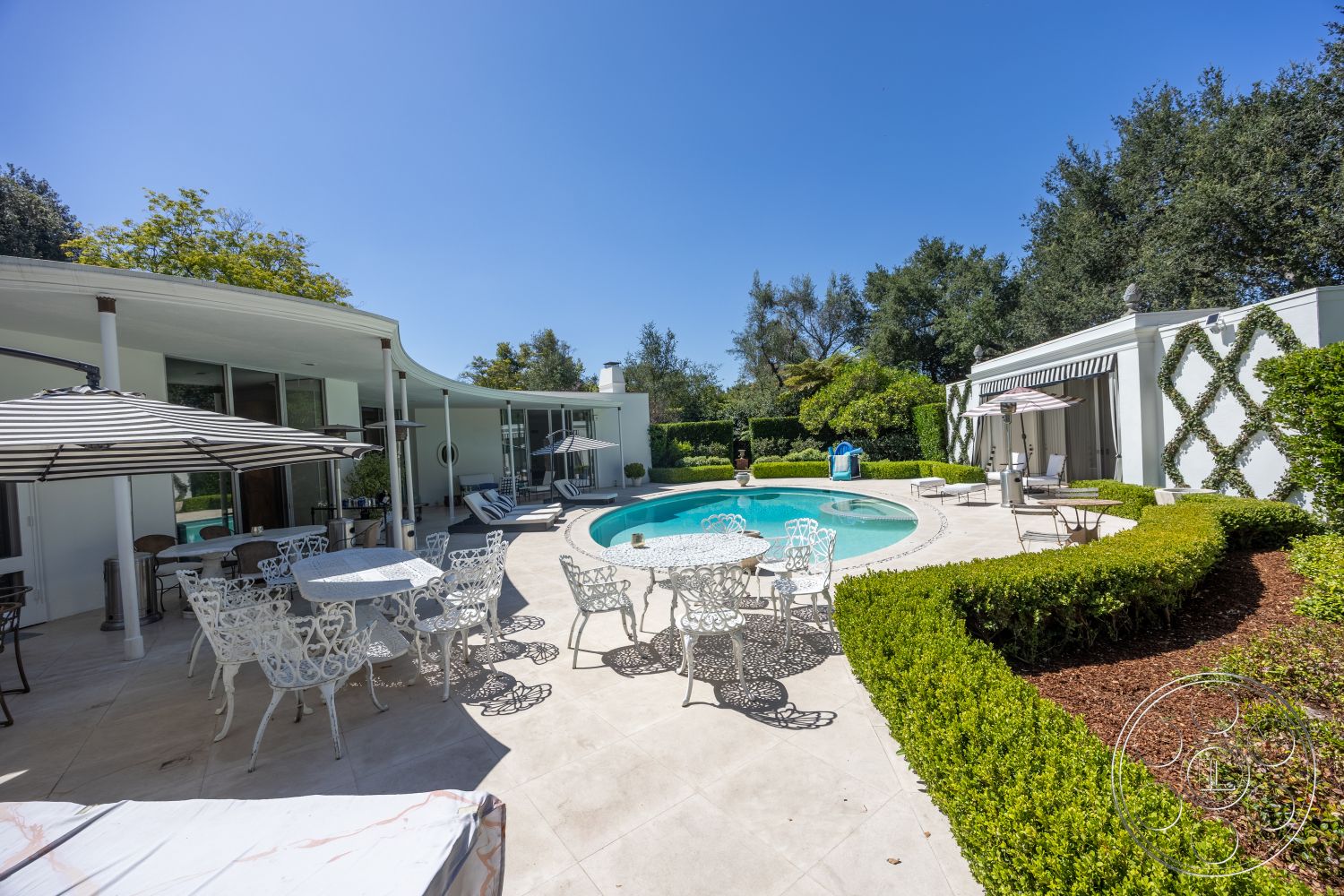 Retro 295 - exterior, landscaped garden, white stucco walls, glass sliding doors, modern architectural style, bush landscaping, umbrella shading, metal patio furniture, sunny outdoor setting, circular pool, concrete patio flooring, brick garden borders, wood garden accents, curved building design, in-ground pool area