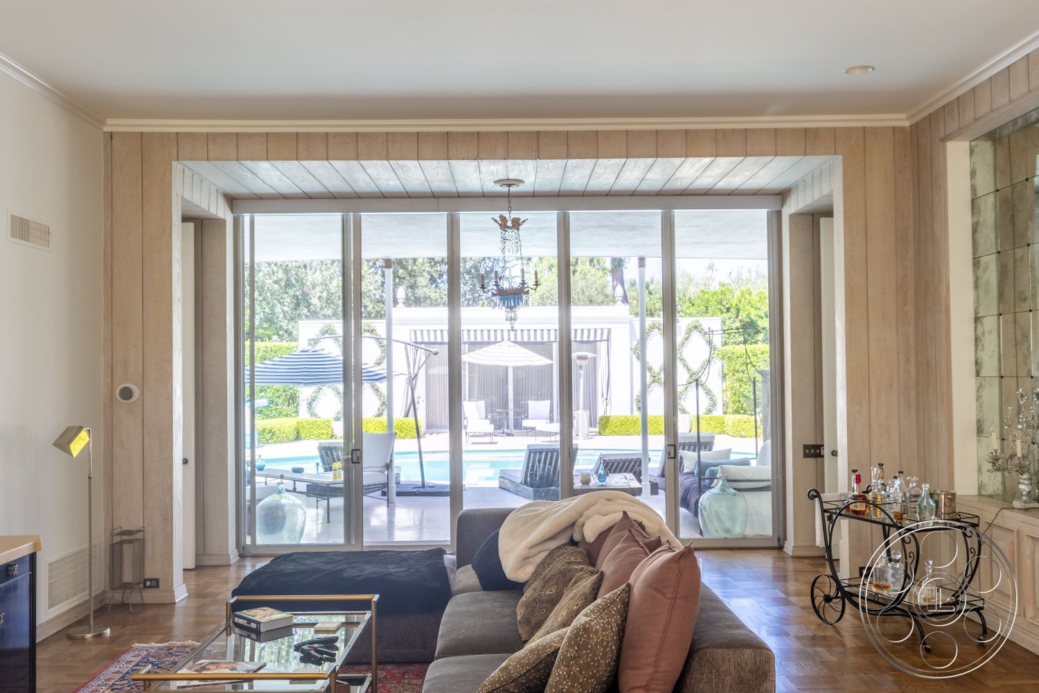 Retro 295 - neutral color palette, decorative area rug, glass sliding doors, modern coffee table, floor-to-ceiling windows, vaulted ceiling, textured wall finishes, open living space, contemporary chandelier, hardwood living room floors, bar cart with glassware, light wood panel wall treatment, beige sofa with patterned cushions, indoor-outdoor flow to pool area