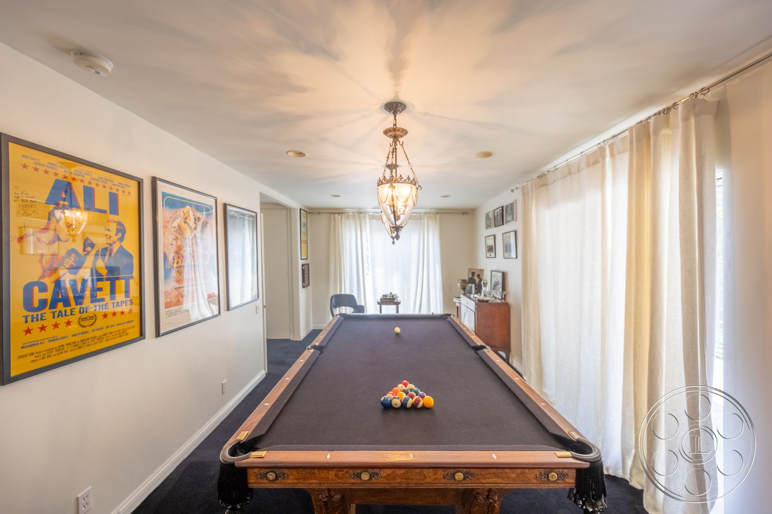 Retro 295 - decorative wall art, laminate flooring, white sheer curtains, glass chandelier lighting, pool table room, soft beige painted walls, dark wood pool table, black felt pool table surface, wooden side cabinet