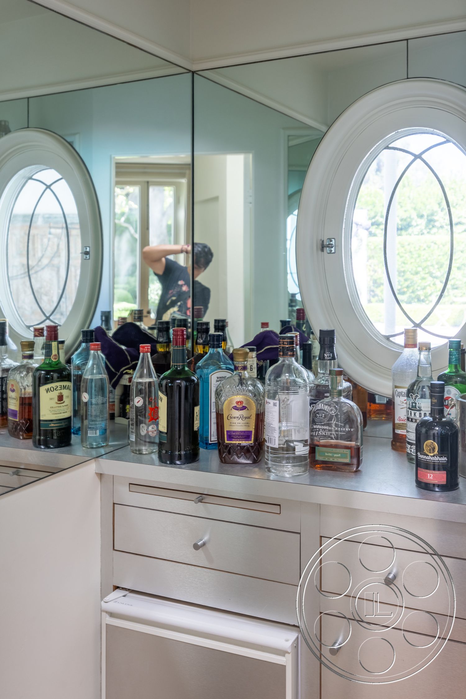 Retro 295 - mirrored wall, modern minimalist style, stainless steel bar countertop, round window with grid design, sleek cabinetry with brushed metal handles, refrigerated wine cooler, display of various liquor bottles
