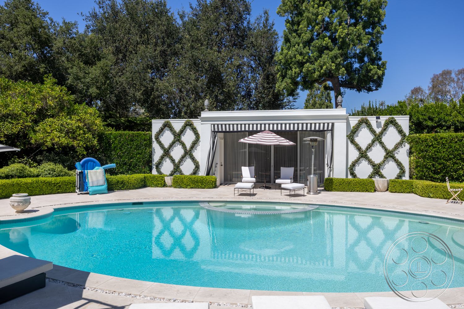 Retro 295 - exterior, white stucco walls, glass sliding doors, concrete pool deck, intimate outdoor space, stone planter, poolside lounging area, outdoor heater, potted plant decor, oval pool, landscaped garden area, bright sunny setting, modern outdoor lounge chairs, paved patio flooring, green hedge boundary, fixed awning with stripe design, lush surrounding trees
