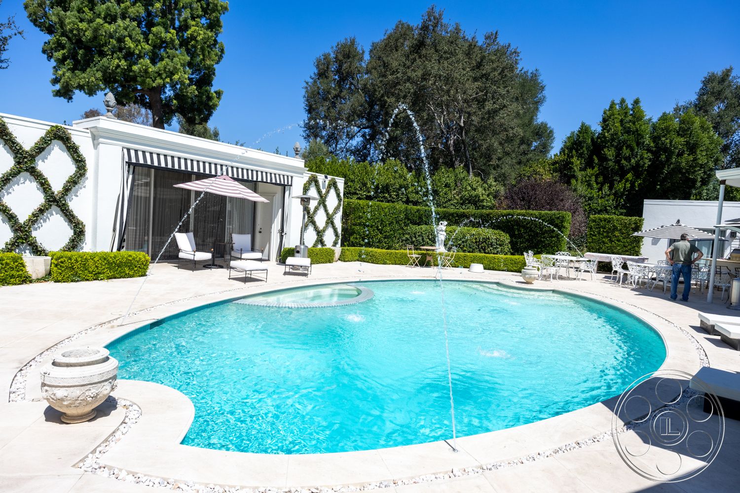 Retro 295 - exterior, glass sliding doors, outdoor lounge chairs, white stucco wall, decorative fountain, oval swimming pool, concrete pool border, natural stone pool deck, sunny backyard setting, garden patio area, landscaped garden with hedge, striped fabric awning, ornate stone planters