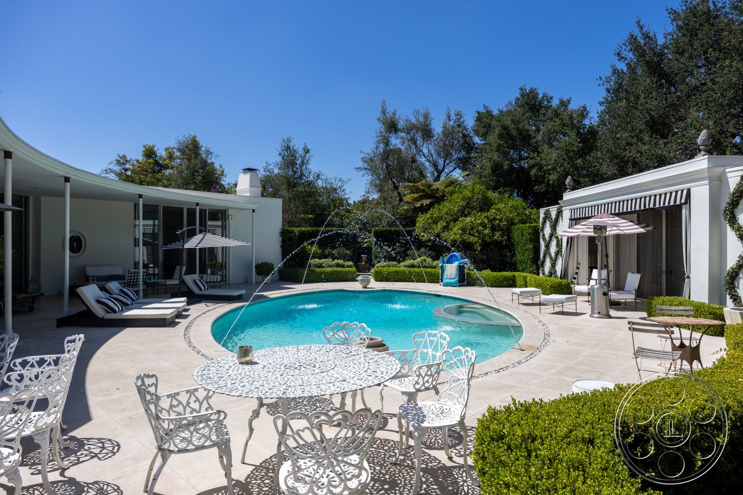 Retro 295 - exterior, landscaped garden, striped umbrella, concrete pool deck, outdoor water feature, oval pool, stone pool border, tile outdoor flooring, sunlight casting shadows, patio lounge area, white metal patio furniture, curved modern home design, evergreen shrubbery