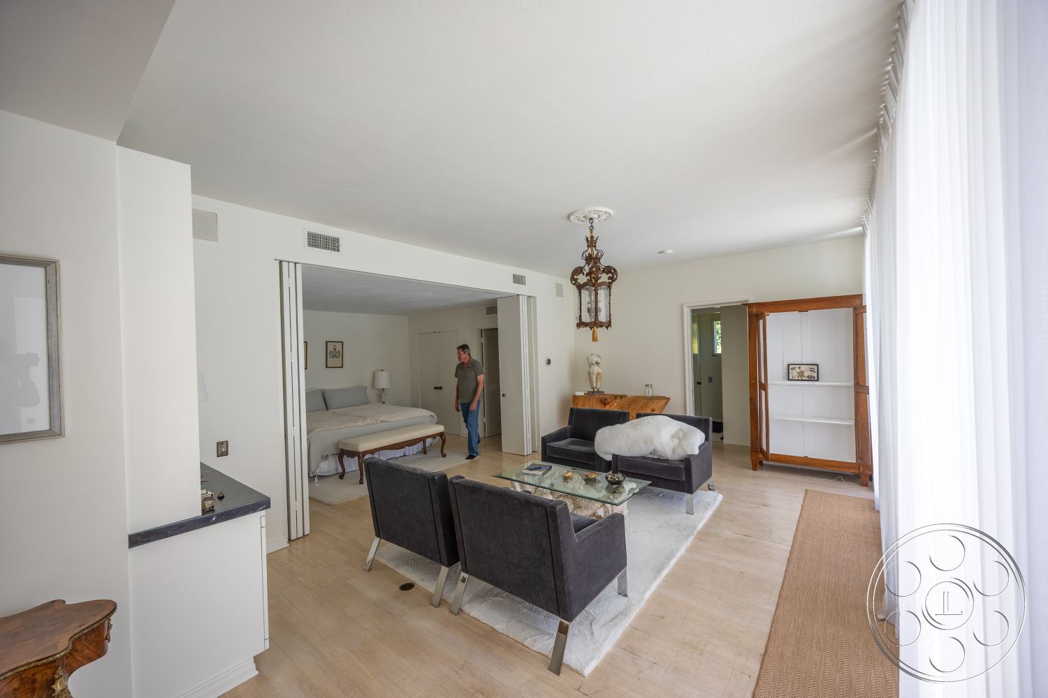 Retro 295 - minimalist decor, neutral color palette, hardwood flooring, glass coffee table, wooden side table, white painted walls, coffered ceiling details, chandelier lighting fixture, light hardwood living room floors, large windows with sheer white curtains, built-in wooden shelving unit, black upholstered armchairs, bedroom area with grey bedding, sliding partition doors